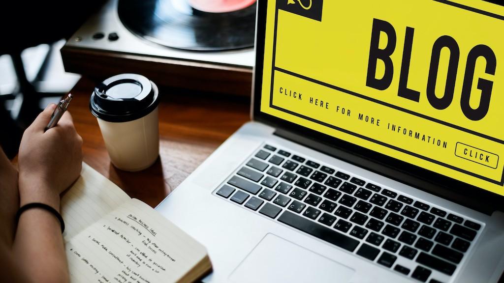 A laptop monitor showing the word BLOG with someone writing blogging ideas on a notebook beside it